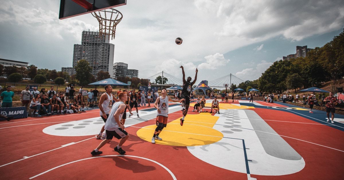 Sportaši igraju košarku na crvenom sportskom terenu.